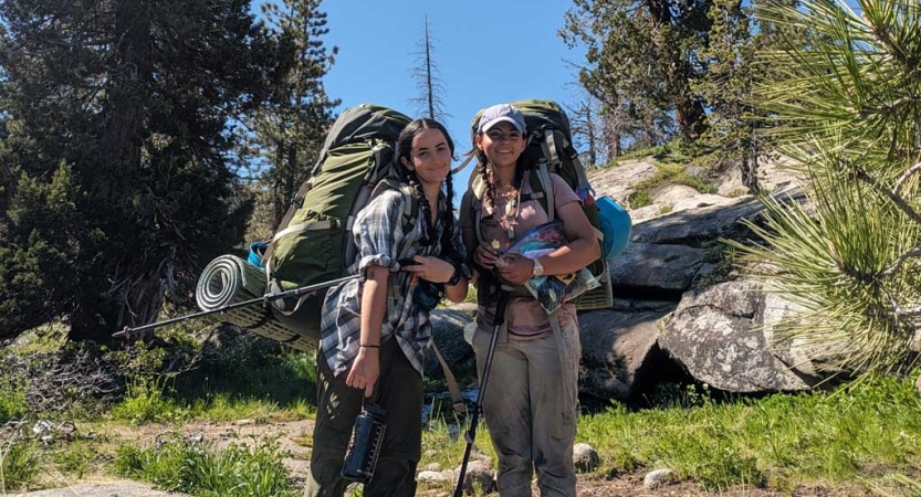 Two people wearing backpacks stand amongst trees and boulders and smile for the photo. 